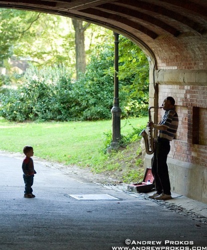 A street scene photo of a child watching intently as a saxophonist plays his horn in Central Park, NYC