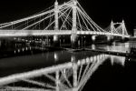 A black and white photo of the lights of the Victorian-era Albert Bridge in London reflected in the waters of the Thames River at night. Limited edition fine art prints are available up to 80 inches in width. 