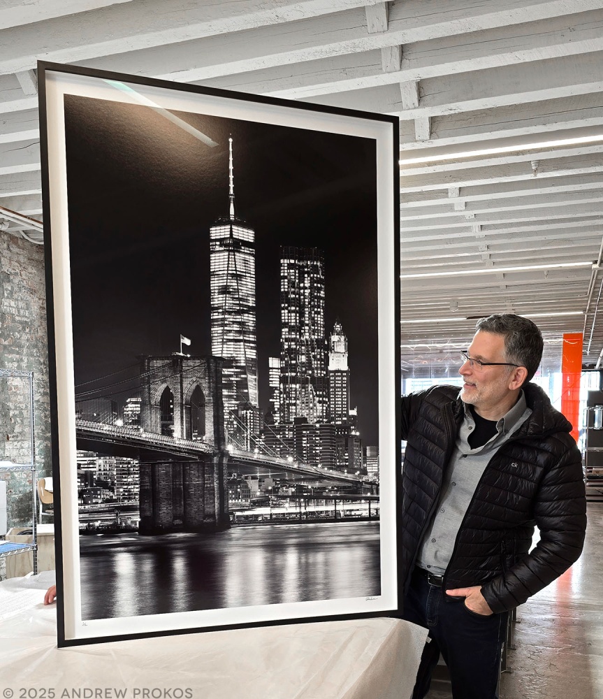 Photographer Andrew Prokos with a framed 60x40 inch black and white fine art print of the Brooklyn Bridge and towers of Lower Manhattan at night.