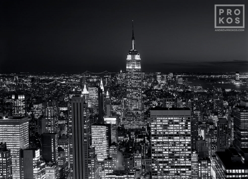 A black and white cityscape photo of the Empire State Building and Midtown Manhattan at night as seen from Rockefeller Center