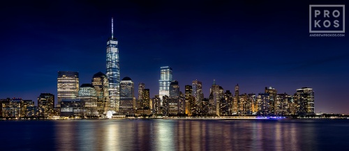 A long-exposure panoramic skyline photo of Lower Manhattan, the Hudson River, and the World trade Center at night as seen from New Jersey.