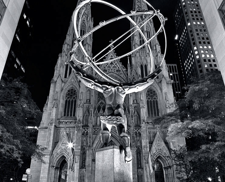 A black and white fine art photo of the Atlas statue at Rockefeller Center and St. Patrick's Cathedral at night, New York City.
