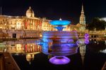 A panoramic photograph of the colorful illuminated fountains of Trafalgar Square, London, at night. Large-format fine art prints of this photo are available up to 120 inches.